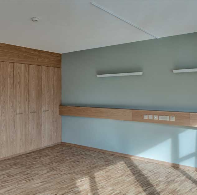 **Alters- und Pflegheim Abendfrieden, Wohnen & Pflege, Kreuzlingen, Thurgau** Die Wand am Kopfende der Betten in den Bewohnerzimmer ist in einem dezenten, eher dunkel gehaltenen Farbton passen zu den Holzoberflächen gestrichen. Die Zimmerbeleuchtung erfolgt über Wandleuchten von Schätti, Modell Andar Care, über den Bettenden mit einem dimmbaren und einzeln verstellbaren Anteil an direktem und inderektem Licht. Der Parkettboden ist in Eiche, geölt. Entgegen dem mit weissen Pigmenten aufgehellten Parkettboden der Korridore ist das Parkett der Zimmer in Natur gehalten und geölt. Die Decken der Privatzimmer sind verputzt.