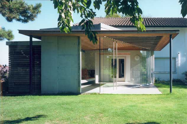 **Gartenpavillon Altnau Thurgau**  Der Gartenpavillon wird als saisonale Erweiterung eines Einfamilienhaus genutzt. Er ist neben den Wohnraum situiert und kann von diesem sowie von der Küche und dem Essraum aus erreicht werden. Die Bodenplatte und die Rückwand sind aus Sichtbeton, das Dach ist einen Holzelementkonstruktion. Grossformatige, rahmenlose Glasscheiben trennen das Innere des Pavillon vom Garten ab, lassen aber den Innenraum mit dem Gartenraum verschmelzen.