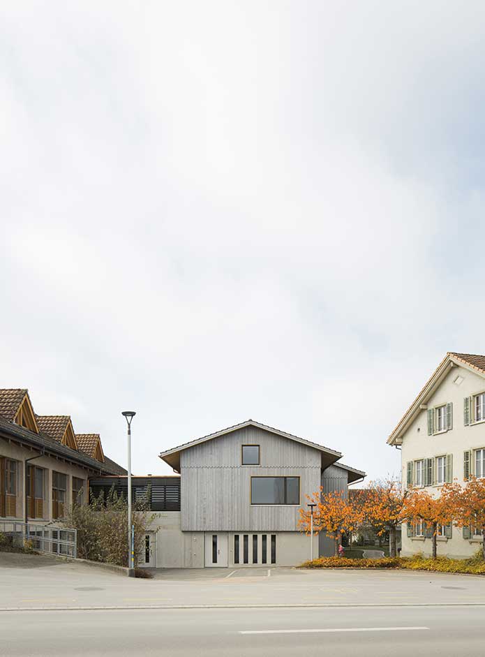 **Erweiterung Stiftung Egnach, Egnach TG, Anlieferung** Der Neubau fügt sich zwischen die bestehende Werkstatt und zwei Wohnhäuser der Stiftung ein. Er übernimmt die Volumetrie und die Dachform des Bestandes und führt die Anlage kammartig fort.  Zusammen mit dem Werkstattgebäude und dem Wohnhaus bildet der Neubau zur Bucherstrasse einen räumlich gefassten Zugangsbereich, welcher für die Anlieferung und Parkierung genutzt wird. Der kleine Hof zwischen Alt- und Neubau dient als Pausen- und Aufenthaltsraum. Ein einfaches Wegnetz verbindet das Werkstattgebäude mit den Wohnhäusern. Eine neue Baumgruppe gliedert den Freiraum und spendet kühlenden Schatten. Der bestehende Rundweg wird um den Neubau herum verlängert. Vor dem Untergeschoss des Neubaus ist ein befestigter Platz vorgesehen, welcher für Arbeiten im Freien genutzt werden kann.  Der neue Arbeitsraum im Erdgeschoss ist barrierefrei an den Bestand angebunden. Die Garderoben sind dem Arbeitsraum vorgelagert. Der Korridor weist eine zum Manövrieren mit Paletten ausreichende Breite auf. Im Untergeschoss unmittelbar beim Eingang ist die Logistik zentral zwischen Eingang und Aufzug und je ein Büro für den Bereichsleiter und den Springer angeordnet. Das Warenlager und der Materialraum sind in wenigen Schritten vom Aufzug aus zu erreichen. Beide Räume verfügen über eine gute Tageslichtsituation und sind so konzipiert, dass sie auch als Arbeitsort genutzt werden können.  Der neue Arbeitsraum ist für eine optimale Nutzungsflexibilität stützenfrei überspannt und reicht bis unter den First. Die Decke über dem Untergeschoss ist zur Reduktion ihrer Aufbauhöhe in der Gebäudemitte auf einer Traglinie aufgelagert und zur Aufnahme der geforderten Nutzlasten in Beton gehalten.  Die Statik - Fundamente, Wände und Decke - ist so dimensioniert, dass eine nachträgliche Aufstockung ohne Verstärkungen aufgenommen werden kann. Das Satteldach würde für die Aufstockung abgebaut und ein Geschoss höher, über einer neuen Zwischendecke, wieder aufgebaut werden. Das Gebäude wäre damit für ein weiteres Geschoss bereit.
