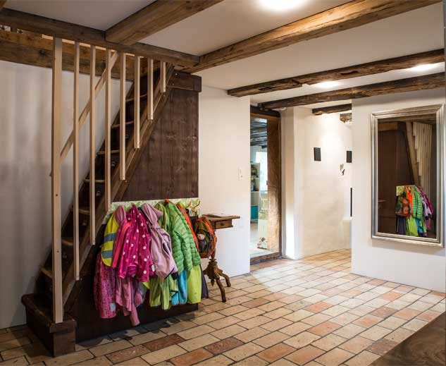 **Entrée und Garderobe im Taunerhaus Hörhausen** Das Wohnhaus wird auch nach dem Umbau mit Anbau über den haupteingang im Altbau betreten. Das Entrée wurde in den ehemaligen Kleintierstall erweitert. Anstelle der ehemaligen offenen Küche mit offener Abzugshutte ist heute die Waschküche und eine Gästetoilette eingebaut worden. Das Entrée führt in das Schlafzimmer der Eltern, die ehemalige Stube mit dem Kachelofen oder in den Anbau, den eigentlichen Wohnteil. Eine steile Holztreppe mit Schubladen in den Stufen, eine Schubladentreppe, für in die Schlafzimmer oder Kinderzimmer im Obergeschoss des Altbaus. Der Bodenbelag des Altbaus wurde mit aufgeschnittenen Tonplatten aus alten Ziegelsteinen geplättelt.