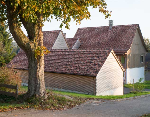 **Die Häusergruppe des neuen Taunerhauses Hörhausen mit der Garage im Vordergrund** Der aus drei Teilhäuser mit je einem eigenen Giebel bestehenden Anbau, die Garage und der Kleintierstall für die Ziegen bilden zusammen einen neue Häusergruppe in der Siedlungsstruktur des Dorfes Hörhausen. An der Natstelle zwischen Altbau und Anbau weden die Volumen und die Dächer miteinander verschnitten. Dieser Übergang ist auch im Innenraum gut lesbar. Alle Dächer sind mit alten Biberschwanzziegel in changierender Färbung gedeckt. Die Fassadenbekeidung der Neubauten sind mit Lärchenholzlatten im Querschnitt von 5 x 5 cm verleidet.