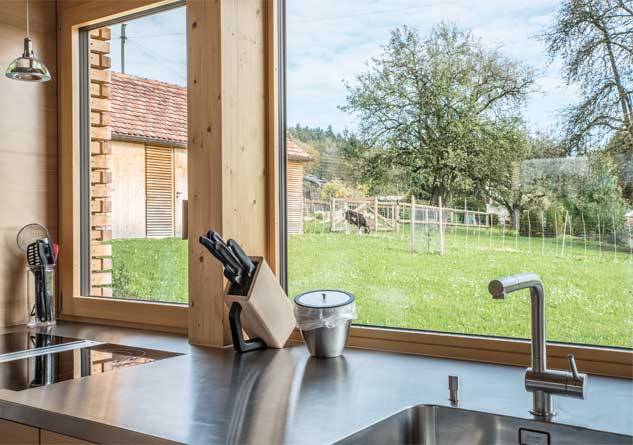 **Der Ausblick aus dem Küchenfenster des Anbaus des Taunerhauses Hörhausen**  Ein grosses Panoramafenster unmittelbar an der Arbeitsplatte der Küche ermöglicht einen weitläufigen Blick in den Landschaftsraum des Gartens und hinüber zum Kleintierstall und den Ziegen.