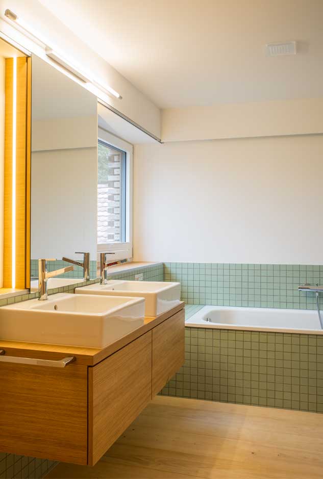 **Badezimmer im Anbaus des Taunerhauses Hörhausen** Im Obergeschoss des zweigeschossigne Neubauteils ist neben einem Schlafzimmer ein Bad eingebaut. Dieses ist über die Galerie auch von den Schlafräumen des Altbaus erreichbar. Es beinhaltet eine Badewanne und eine Dusche sowie zwei Lavabos und ein Toiletten-Nische. Dusch- und Badebereiche sind mit Feinsteinzeugplatten von Winkelmann gefliesst. Der Parkettboden des Galerie ist bis an die Badewanne geführt.