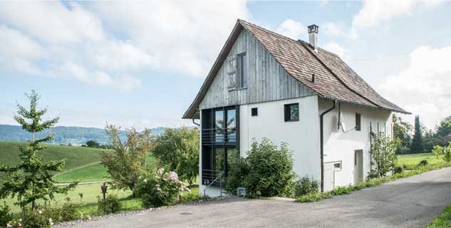 **Ansicht West ** Die Westfassade des Taunerhauses ist mit neuen in Stahlrahmen gefassten Fenster-Öffnungen ausformuliert. Der Klappladen des Dachfensters übernimmt in geschlossenem Zustand die offene Holzschalung des Giebeldreiecks. Die Dachflächen sind mit Isofloc isoliert und mit Bieberschwanz-Ziegeln gedeckt. Der historische Dachabsatz zwischen dem ursprünglichen Wohnteil und dem Ökonomieteil wurde beibehalten.