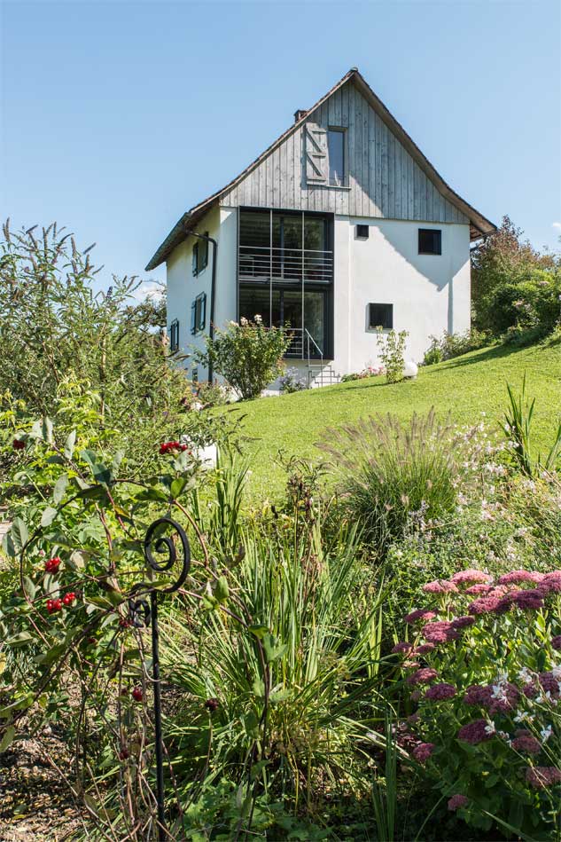 **Ansicht West, Gartenseite ** An der ursprünglich geschlossenen Westseite des Taunerhauses konnten neue, grosszügige Öffnungen zugefügt werden. Ein Stahlrahmen fasst die raumgrossen mit Hebeschiebetüren versehenen Öffnungen über zwei Geschosse zusammen. Ein kleiner Balkon im Obergeschoss ermöglicht einen Austritt aus dem Arbeitszimmer. Die Fenster der Badezimmer und des Schrankraums sind frei verteilt über die zweite Hälfte der Fassade und halten sich in ihrer Positionierung im Gleichgewicht. Der Garten im Vordergrund ist reichhaltig mit einheimischen Blumen und Sträuchern bepflanzt.