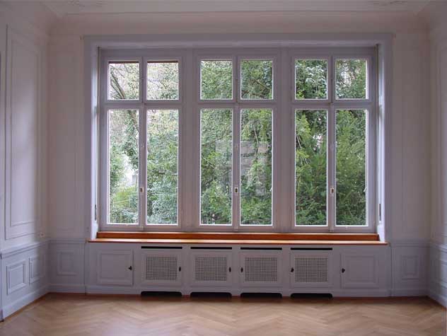 **Fensterfront Hochparterre** Die dreiteilige Fensterfront ist mit Oberlichtfenstern und einem Kämpfer versehen. Handgeschmiedete Stangenbeschläge mit Dreholiven in Messing schliessen die Fenster. Seitlich in den vertäfelten Fensterleibungen befinden sich die Rollgurten für die Holzrollladen. Unter dem Fenstersims in Eiche befindet sich der Rippenradiator aus Grauguss. Dieser wird durch einen mit Zierlöchern versehene Heizkörperverkeidung verdeckt.