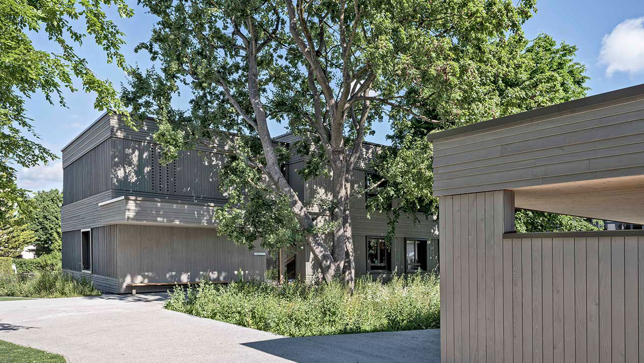 **Kinderhort Tannegg, Kreuzlingen Thurgau** Dem neuen Hortgebäude vorgelagert ist ein grosszügiger Spielhof mit Bauminseln in den Bereichen der bestehenden Bäumen. Diese sind mit Wiesenblumen eingesät und einzelne sind mit einer Sitzbank zum Spielen und Verweilen eingefasst. Der Haupteingang öffent sich direkt in diesen Spielhof. Ein weit  auskragendes Vordach sützt diesen und einen Sitzbank bietet Platz zum Warten am Schatten. Das Gebäude ist ab der Bodenplatte ein reiner Holzbau. Die Bereiche unter und über den Fenstern sind mit einer horizontalen diese zwischen den Fenstern mit einer vertikalen hinterlüfteten Stulpschalung verkleidet. Die Fenster sind mit einem Fenstergewände in Aluminium allseitig eingefasst. Die Bleche sind einbrennlackiert und die Farbe erhält durch den Zusatz von Eisenglimmer eine schimmernde Tiefenwirkung. Kleine Vor- und Rücksprünge im Gebäudevolumen gliedern dieses und reagieren damit auf die nahe Umgebung.